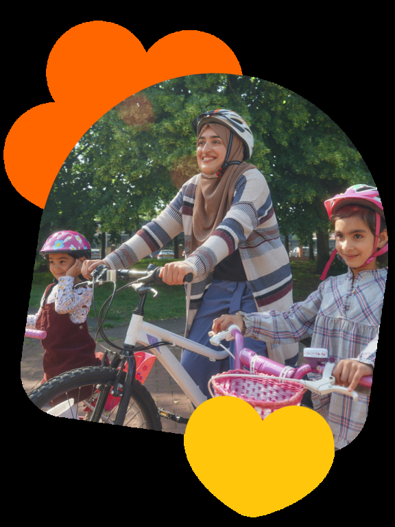 A woman cycles with two children along a park path on a sunny day, with trees in the background.